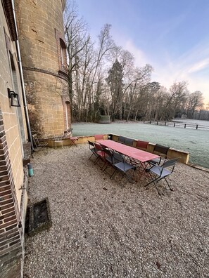 Une terrasse en gravier avec une table et des chaises rouges, un bâtiment en briques et une zone délimitée par une clôture, avec des arbres en arrière-plan.