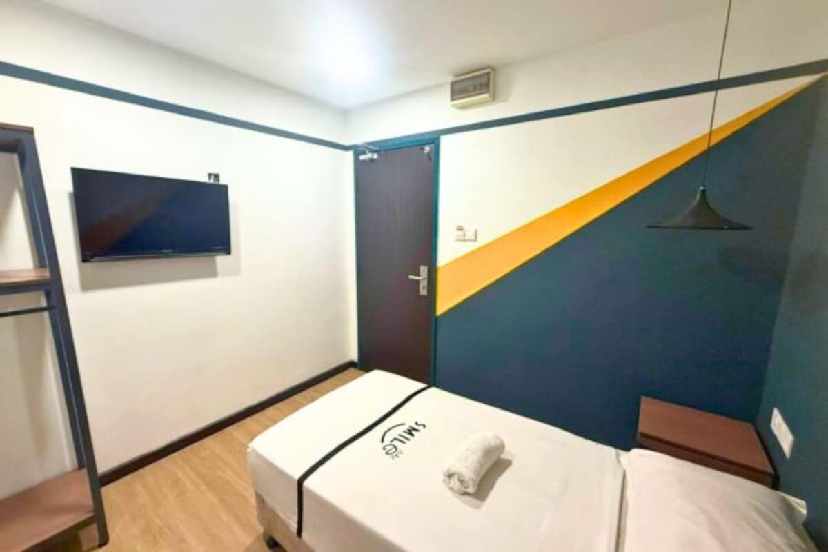 A small, neatly arranged room with a bed, a TV mounted on the wall, and a door.