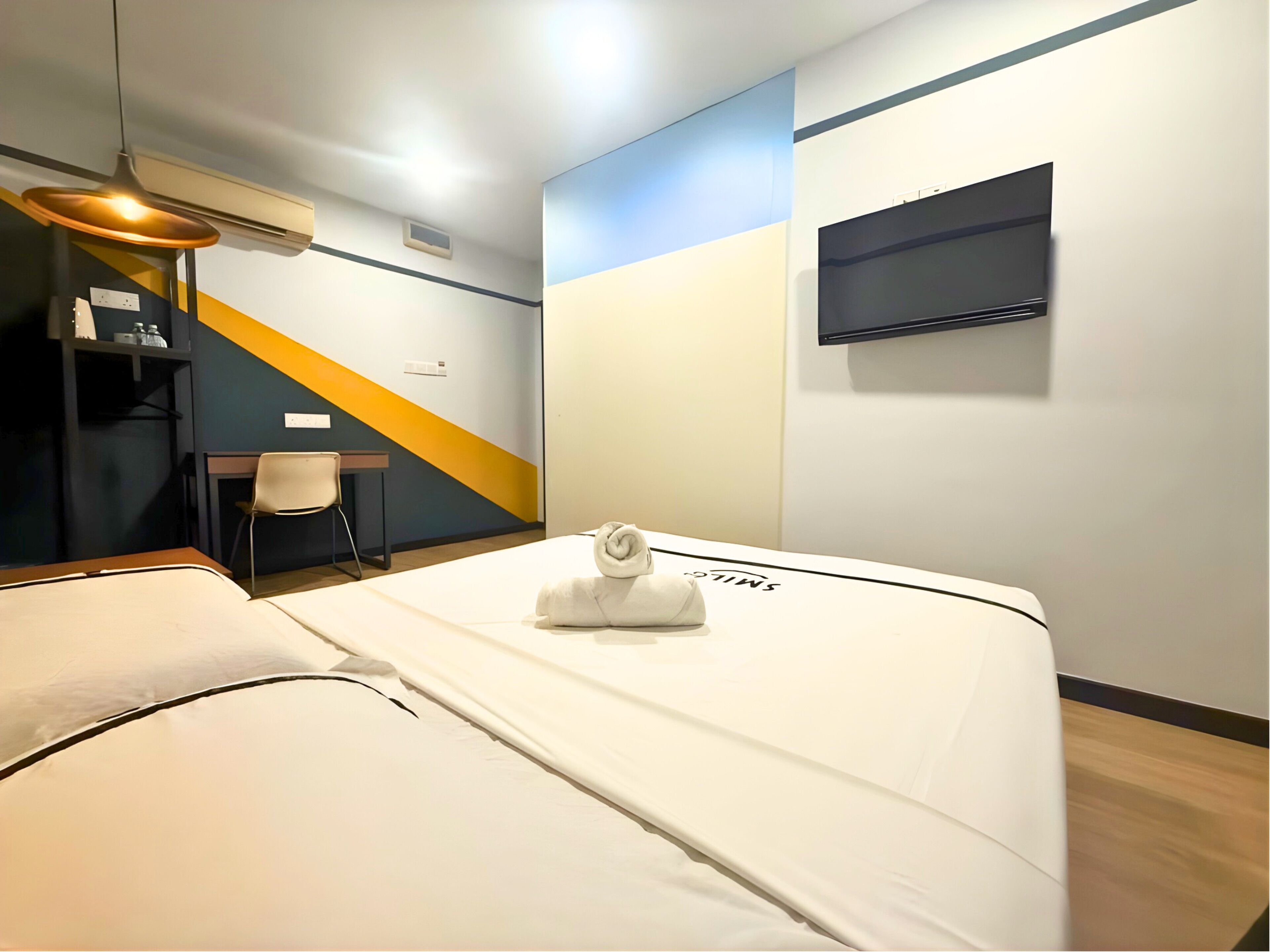 A hotel room with a bed, a desk, a chair, a flat-screen TV mounted on the wall, and an air conditioning unit.