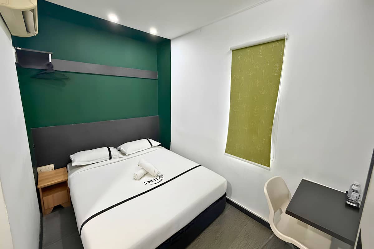 Una camera d'albergo con un letto, una scrivania piccola, una sedia, una finestra con persiana verde e un condizionatore d'aria a parete.