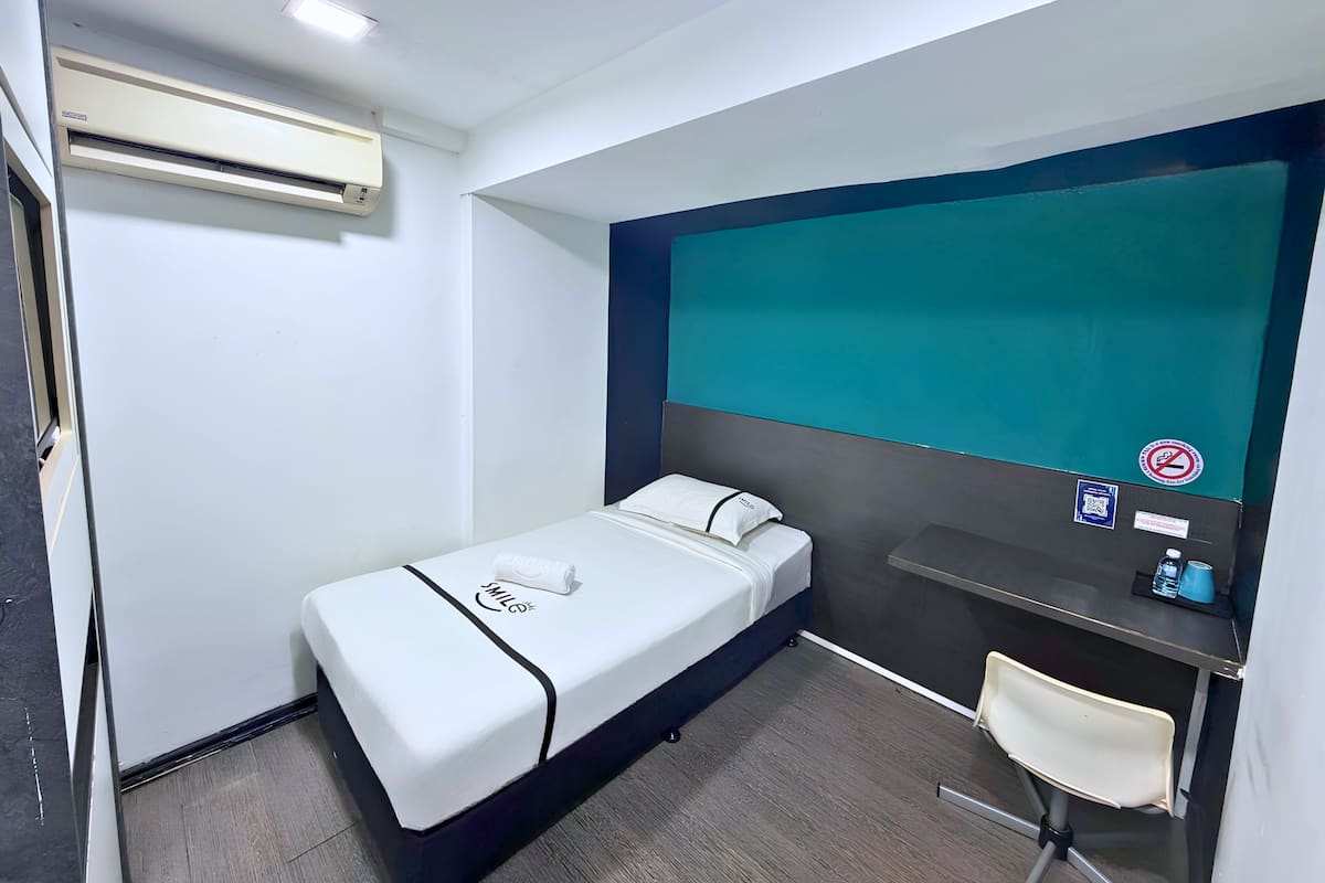Un letto singolo con materasso bianco e rifiniture nere, una piccola scrivania con sedia, una parete color acquamarina e una finestra.
