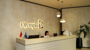 Een receptiebalie met een bordje met daarop 'Olympia Villas & Hotel', vlaggen en een receptiemedewerker.