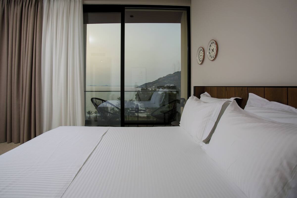 A hotel room with a large bed, a balcony with a view of the sea, and two wall clocks.