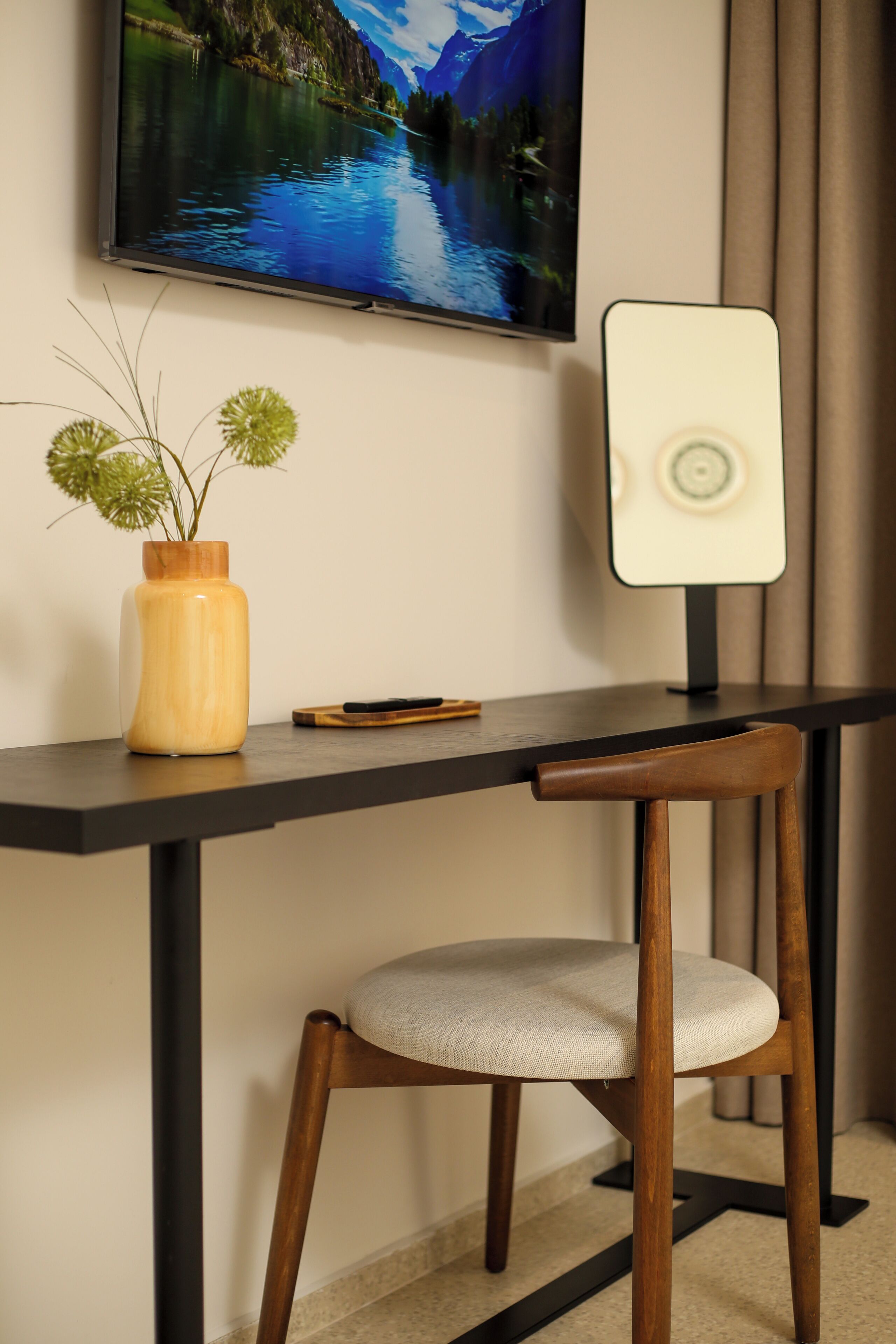 A modern desk with a wooden chair, a vase with green plants, a smartphone, and a TV mounted on the wall displaying a scenic landscape.