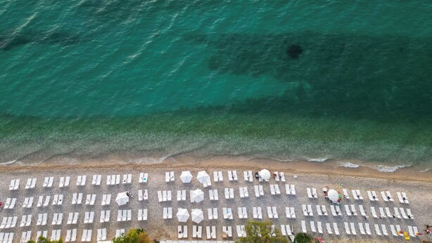 Luchtbeeld van een strand met rijen witte ligbedden en parasols, een heldere turquoise zee en een zandstrand.