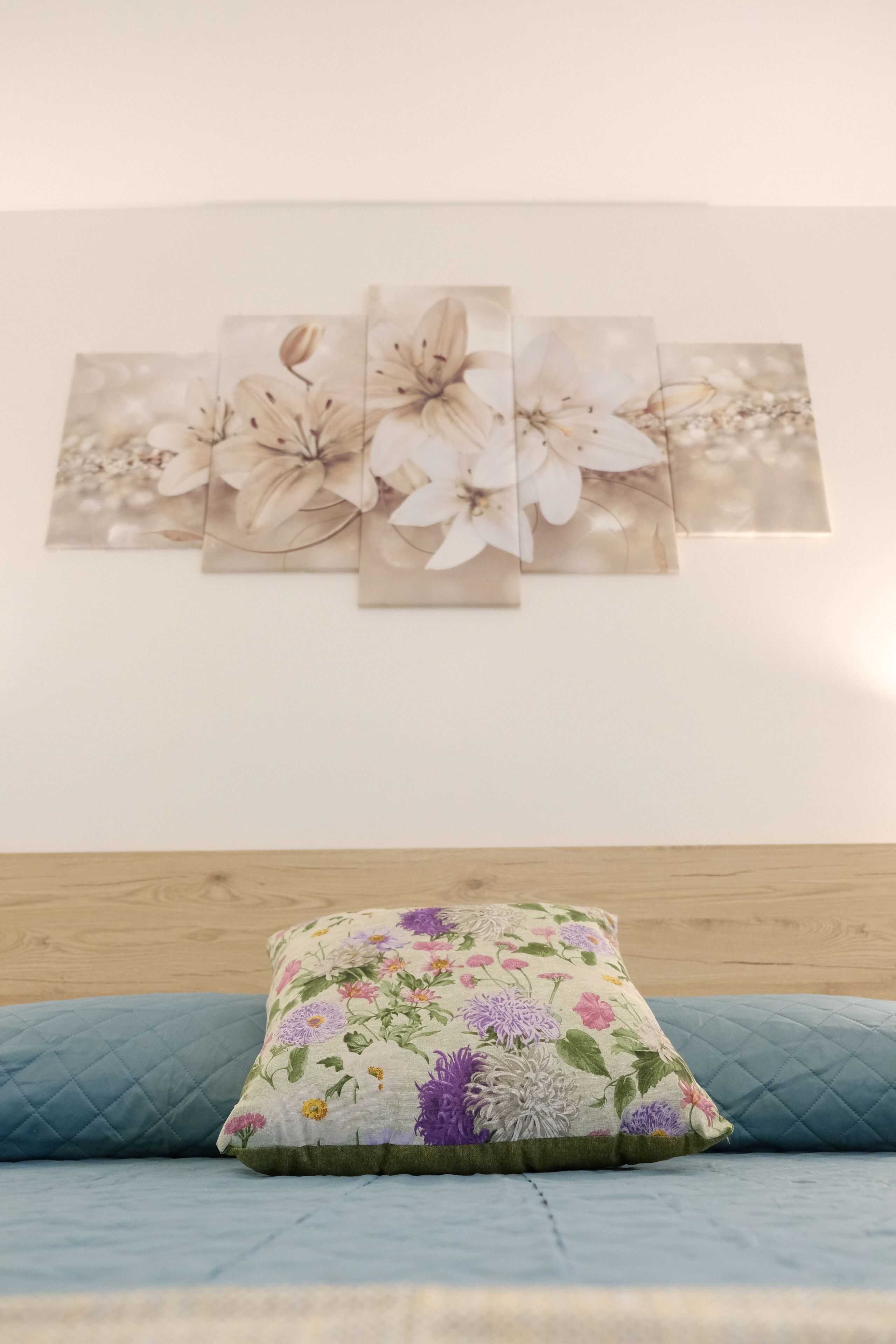 En säng med ett blått, vadderat överdrag, en prydnadskudde med blommönster och en vägg med en tavla som föreställer blommor.
