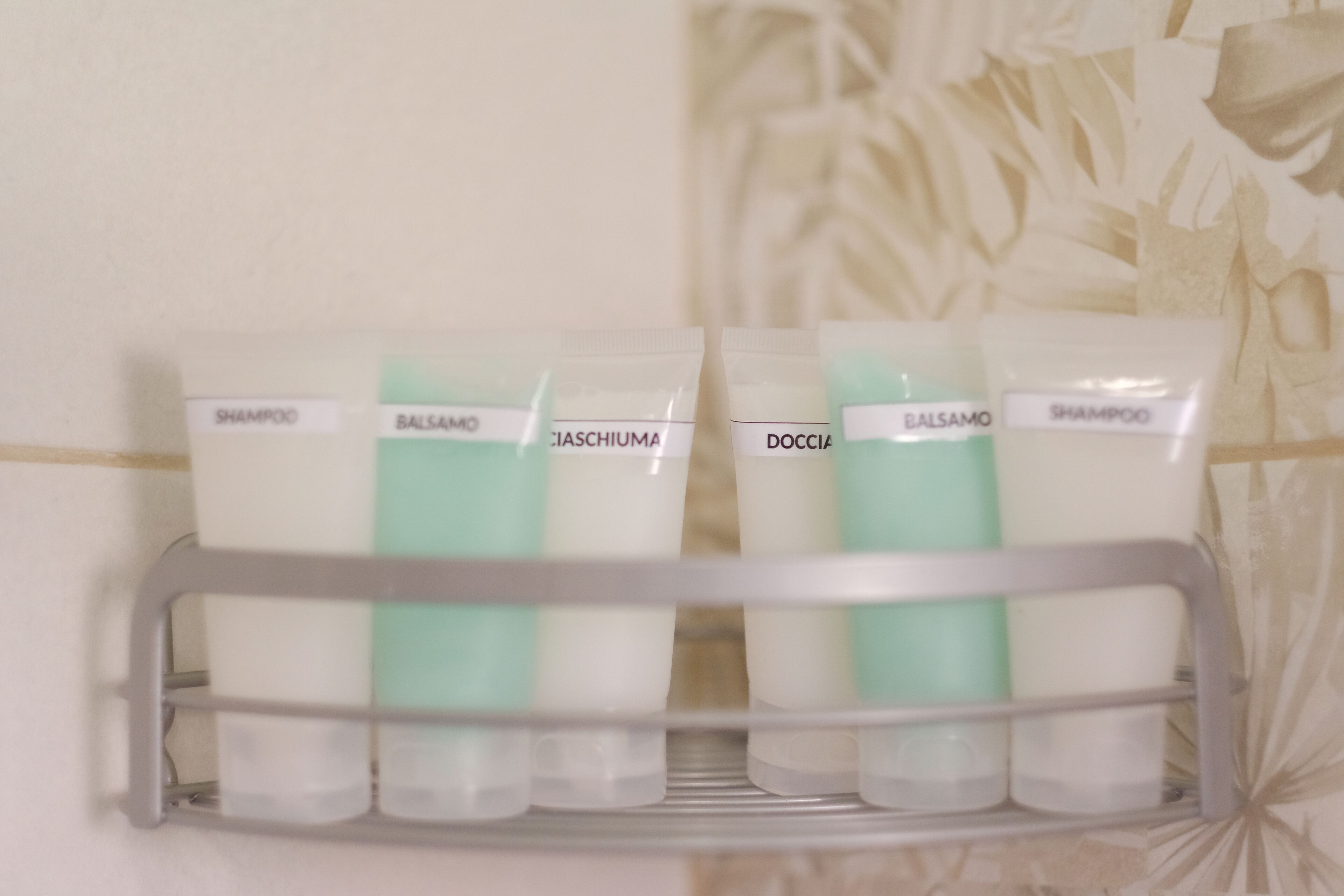 A bathroom shelf with four toiletries: shampoo, balsamo, cicchiuma, and doccia.