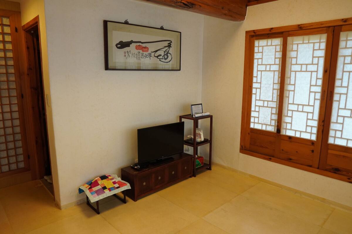 Una habitación con una televisión sobre un mueble de madera, un cuadro en la pared y una ventana con marcos de madera.