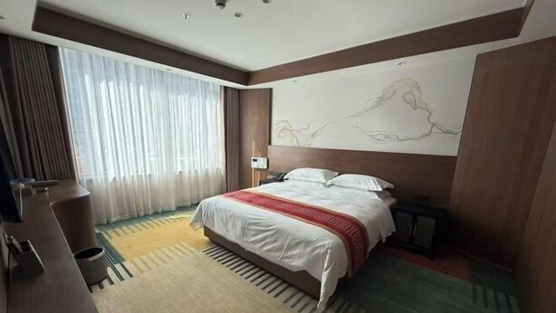Una habitación de hotel con cama, escritorio, silla, ventana grande con cortinas y una obra de arte en la pared.
