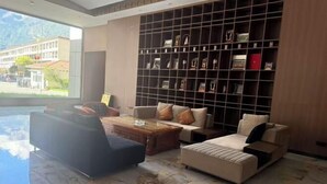 A modern living room with a sofa, armchair, and coffee table, a wall with a grid of framed pictures, and a large window with a view of buildings and greenery. - Linzhi Hotel (Nyingchi)