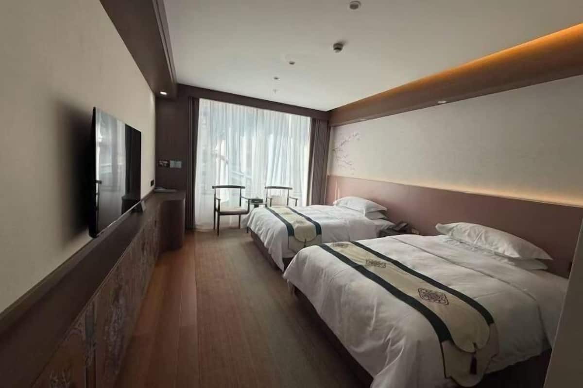 A hotel room with two beds, a flat-screen TV, a chair, and a large window with curtains.