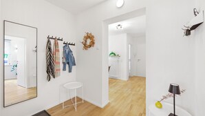 Ein modernes, gut beleuchtetes Zimmer mit einem großen Spiegel, einem Kleiderständer mit Jacken, einem kleinen weißen Hocker und einem Tisch mit Lampe und Blumenvase.