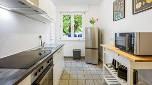 Eine moderne Küche mit Einbauherd, Mikrowelle und Kühlschrank. Ein Fenster bietet Blick auf Bäume, und ein Mülleimer ist vorhanden.
