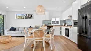 A modern kitchen with white cabinets, a black refrigerator, and a dining area with wicker chairs and a glass table. - Currawong - Coastal Bliss - walk to the beach (Rye)