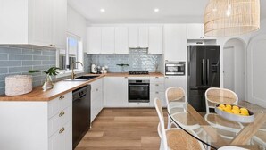 A modern kitchen with white cabinets, a black refrigerator, a stove, and a dining area with wicker chairs and a glass table. - Currawong - Coastal Bliss - walk to the beach (Rye)