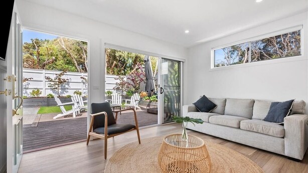 A modern living room with a sofa, a chair, a wicker table, and a view of the outdoors through glass doors. - Currawong - Coastal Bliss - walk to the beach (Rye)