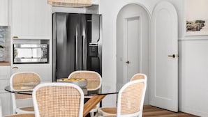 A modern kitchen with white cabinetry, a stainless steel refrigerator, and a dining area with wicker chairs and a glass table. - Currawong - Coastal Bliss - walk to the beach (Rye)