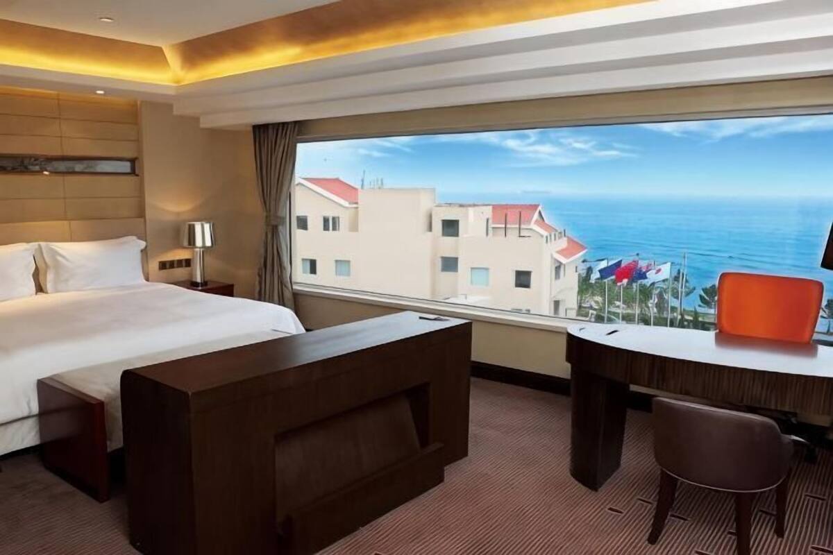 A hotel room with a bed, a desk, a chair, and a large window offering a view of the sea and buildings.