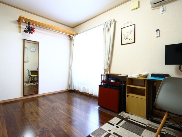 Una habitación con suelo de madera, un escritorio con computadora, una silla, un televisor y una ventana con cortinas.