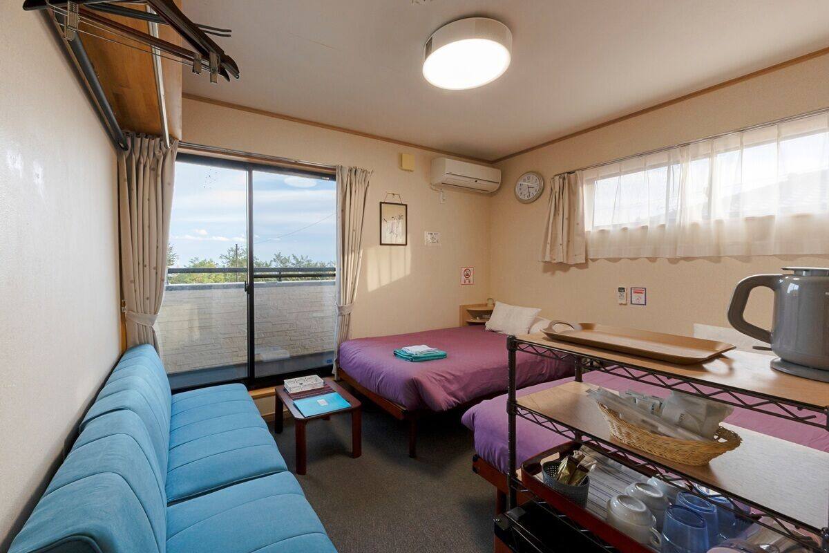 A compact room with a blue sofa, two beds with purple bedding, a small table, and a shelf with items.