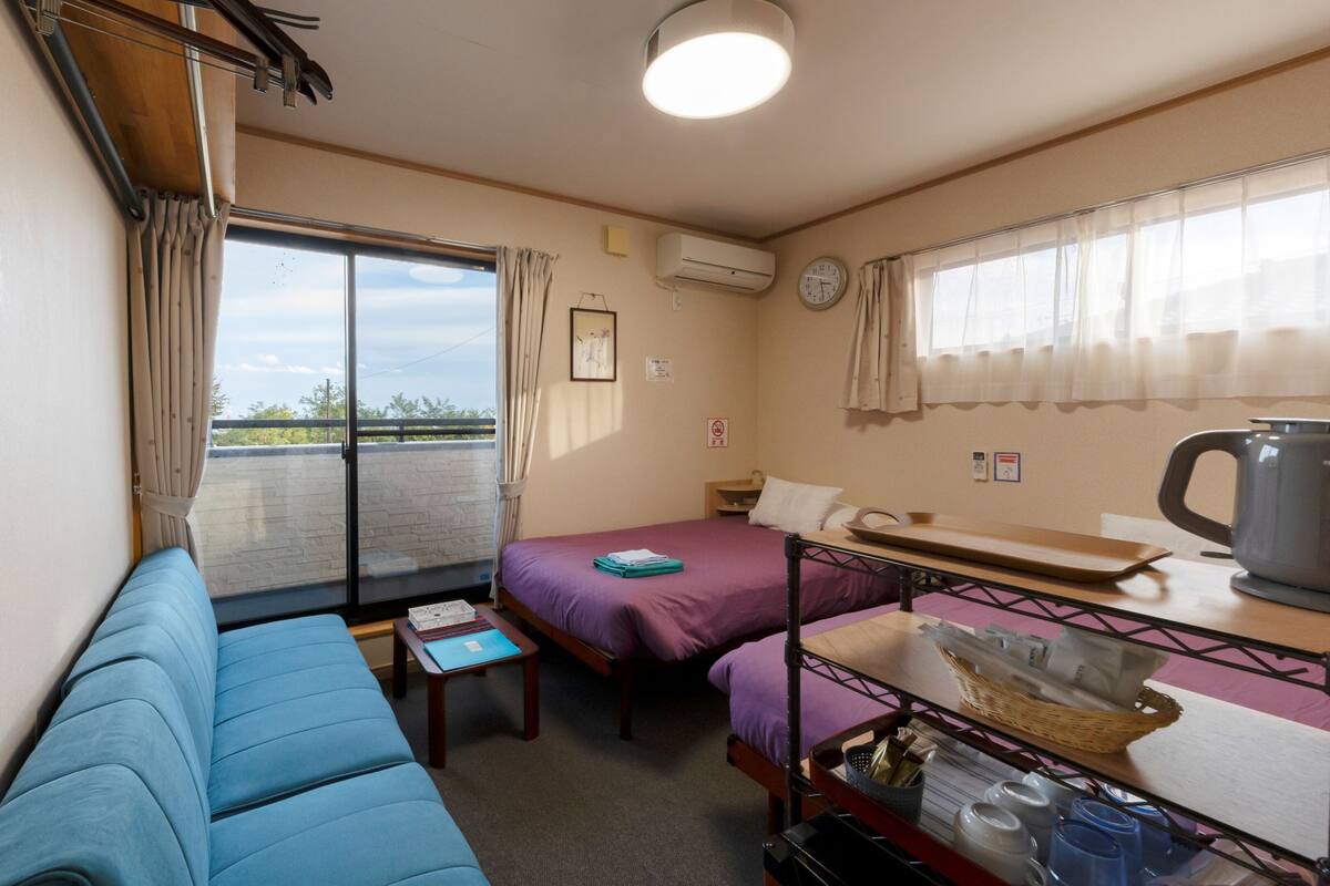 A compact room with a sofa, a bed, a small table, a kettle, and a window with curtains.