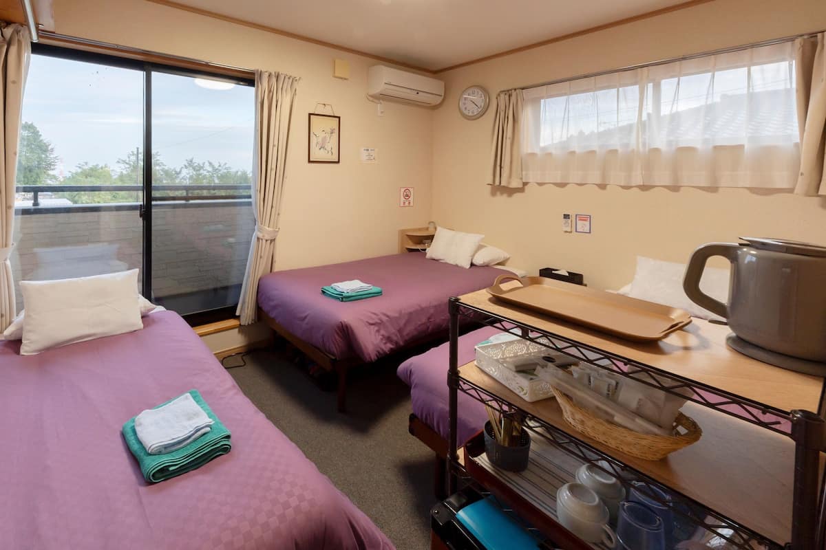A hotel room with two beds, a window with curtains, a clock, an air conditioner, and a small table with a kettle and cups.