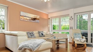 Een woonkamer met een beige bank, een houten salontafel en een schommelstoel. Aan de muur hangt een schilderij en er is een raam met uitzicht op groen.