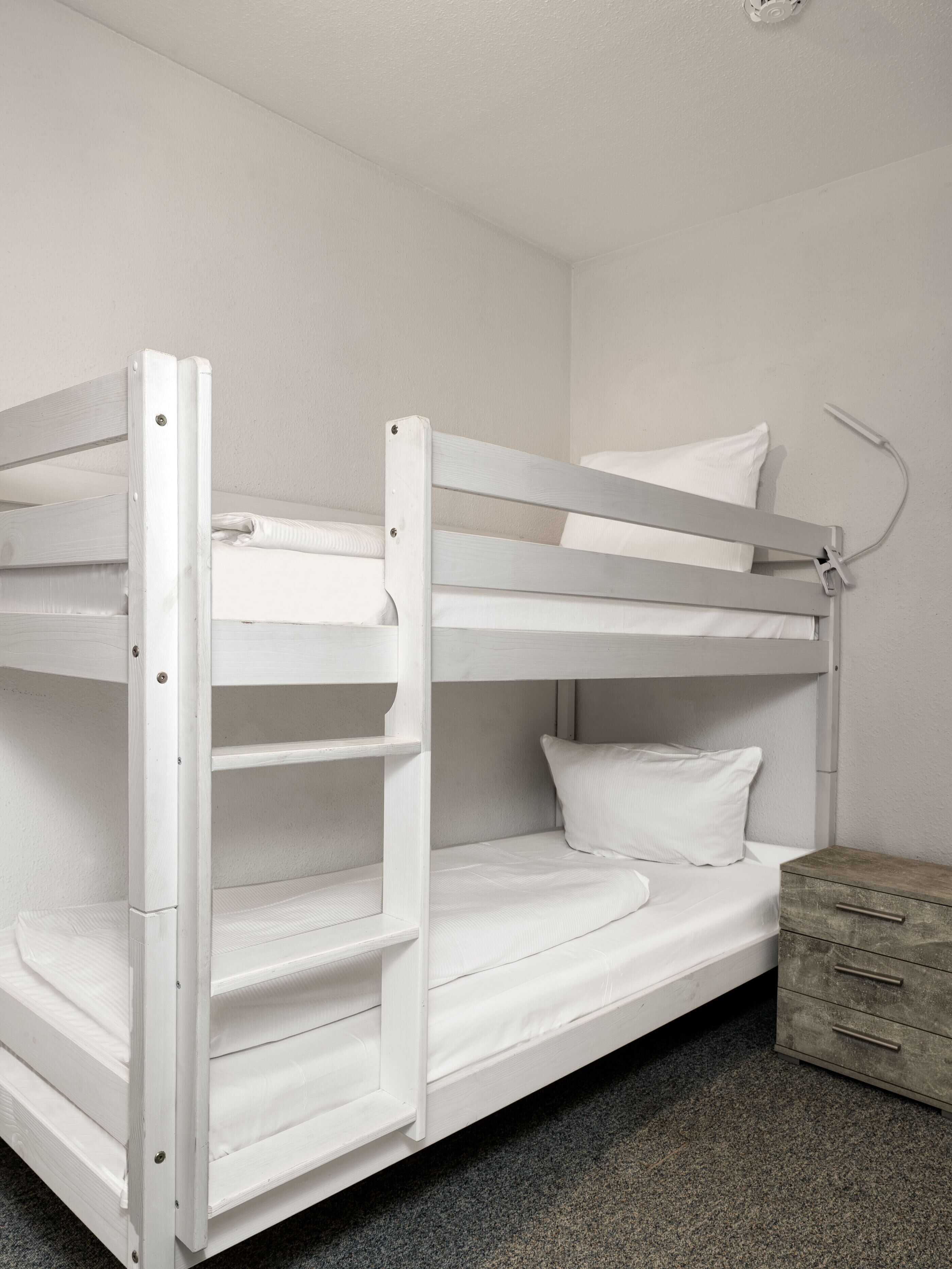 A bunk bed with a wooden frame, a bedside table with drawers, and a wall-mounted lamp.