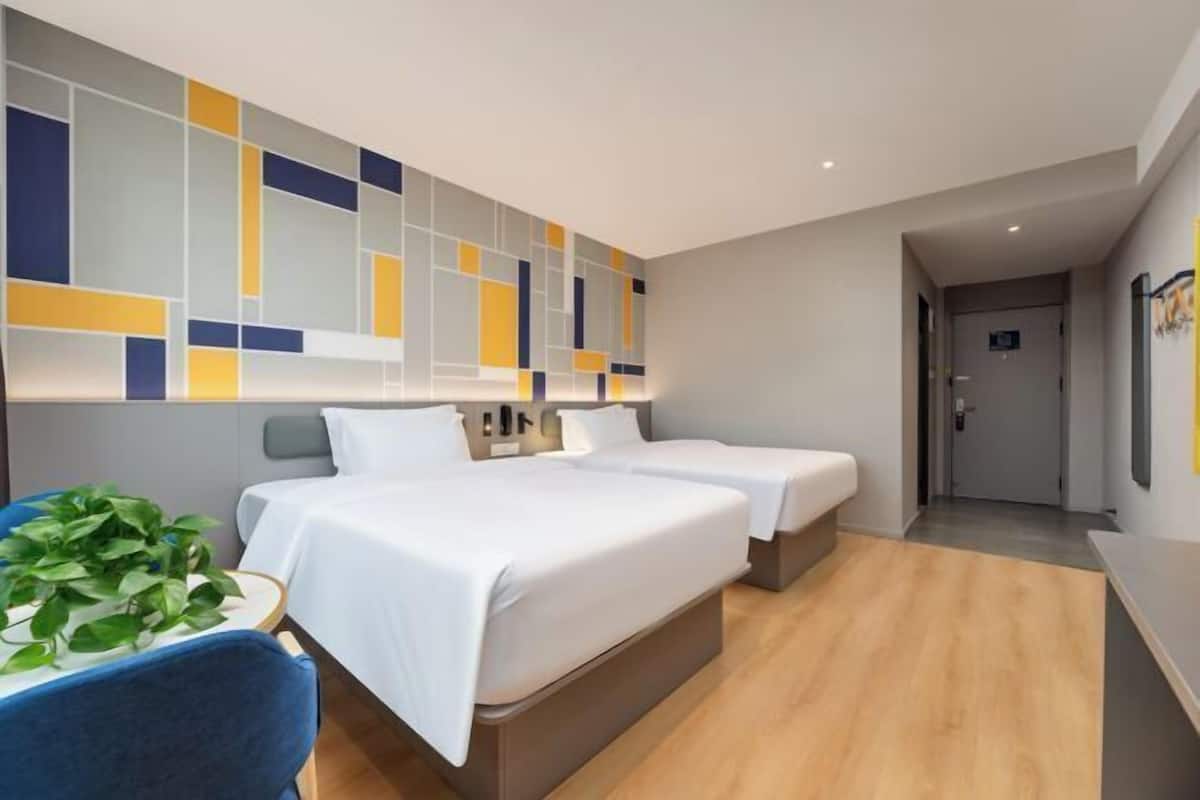 A modern hotel room with two beds, a blue chair, a plant, and a geometric wall design.