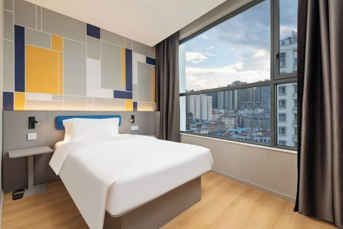 A hotel room with a large window, a bed, a bedside table, and a modern wall design.