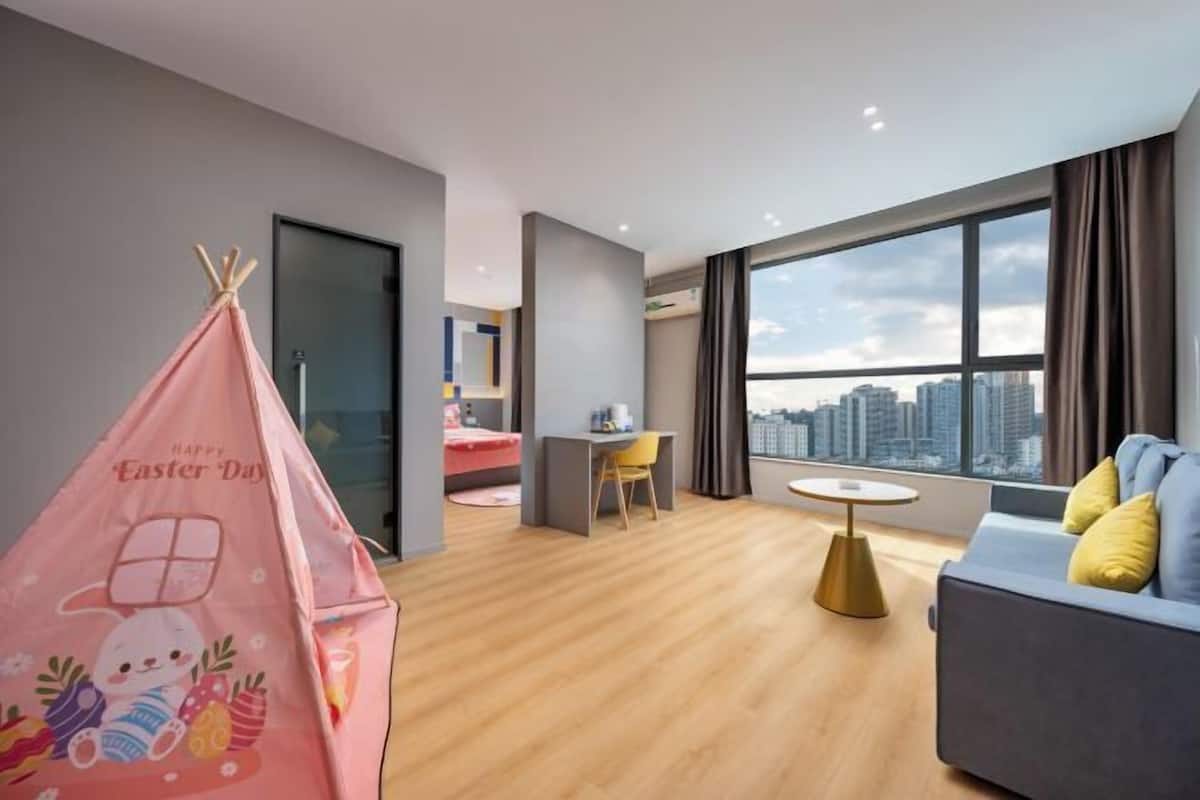 A modern hotel room with a city view, a small desk, a sofa, and a pink Easter-themed tent.