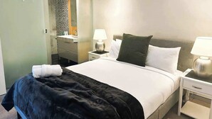 Habitación de hotel con una cama, dos mesitas de noche con lámparas, una mesita auxiliar y un espejo, además se ve un baño a través de una puerta abierta.
