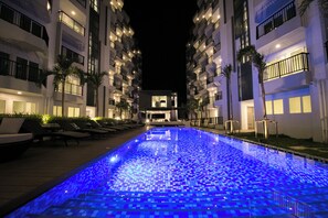 Relax by the illuminated pool surrounded by palm trees and lounge chairs.