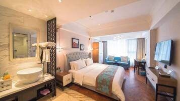 Quarto de hotel com uma cama grande, toucador com pia, televisão e área de estar com sofá e poltrona.