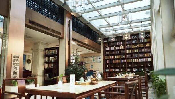 A spacious indoor area with wooden tables and chairs, a large bookshelf, and a glass ceiling allowing natural light.