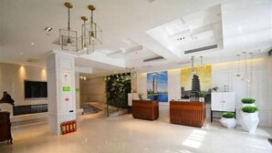 A modern hotel lobby with a reception desk, a large plant, and a painting on the wall.