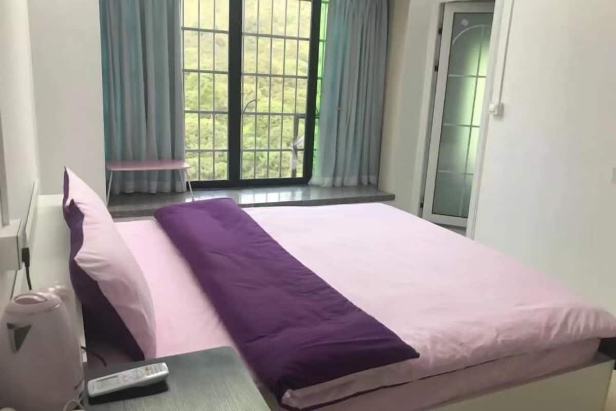 A bed with a purple and pink bedspread, a remote control on the side, and a window with curtains.