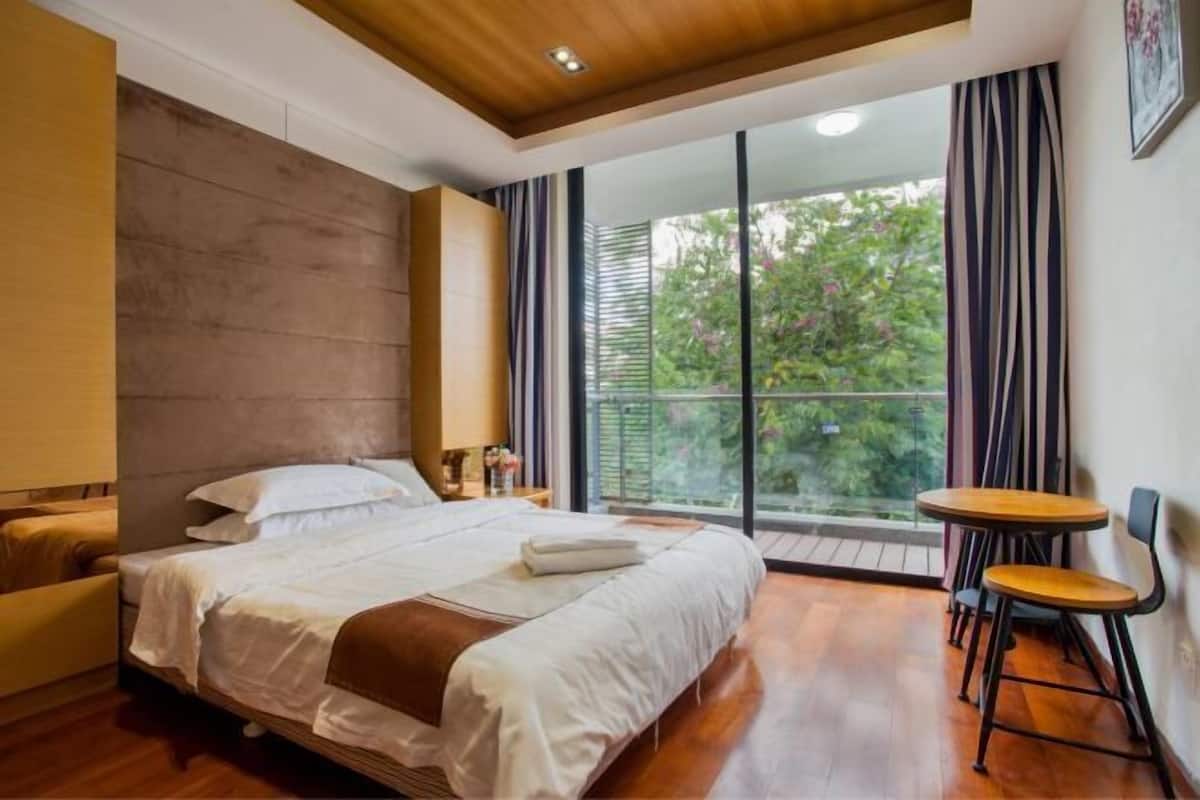 A bedroom with a bed, a small table, and a chair, a window with curtains, and a view of greenery outside.