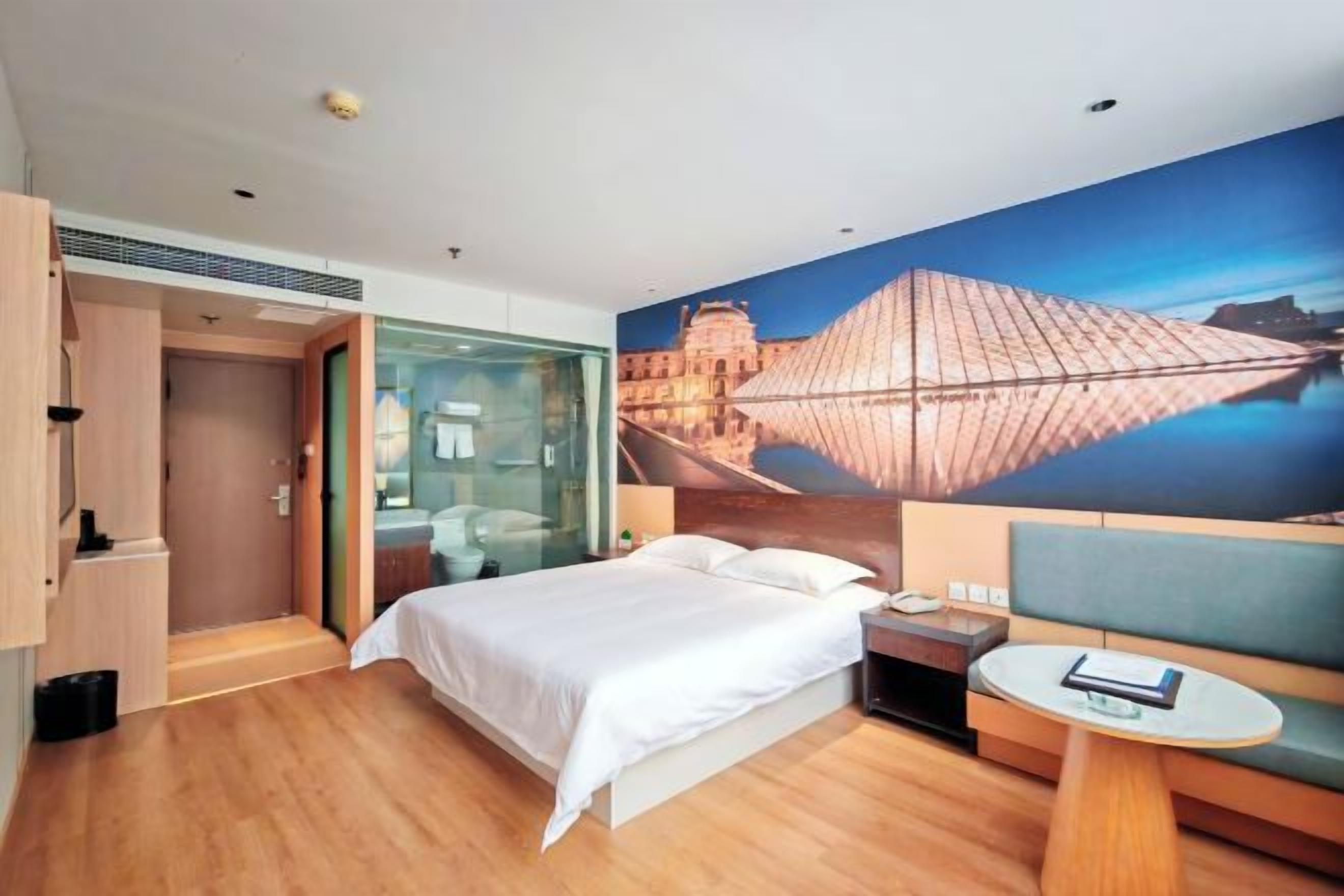 A modern hotel room with a large bed, a glass partition, a small table, and a wall mural.