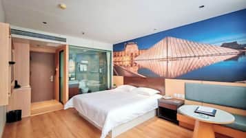 Una habitación de hotel moderna con una cama grande, un separador de vidrio, una mesita pequeña y un mural en la pared.