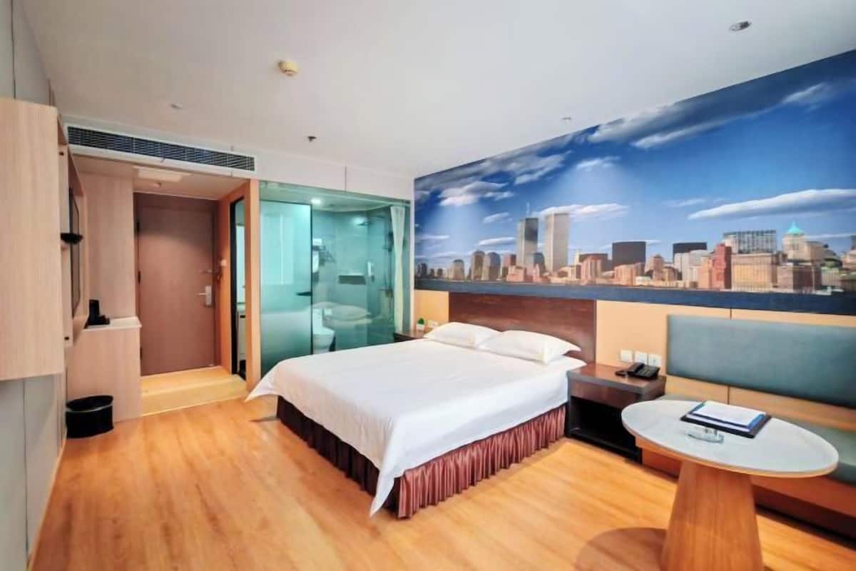 Habitación de hotel con cama, zona de estar con mesa, separador de vidrio y un mural con vistas a la ciudad en la pared.