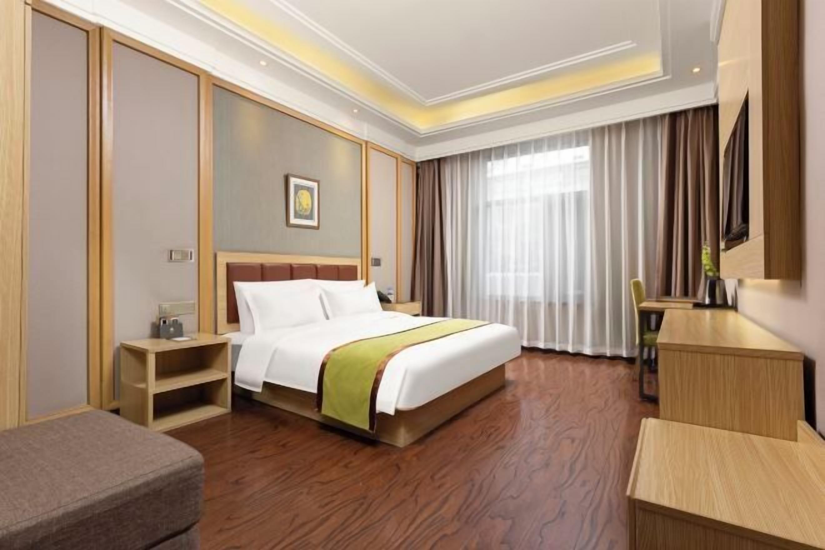 A hotel room with a bed, bedside table, chair, desk, and a large window with curtains.
