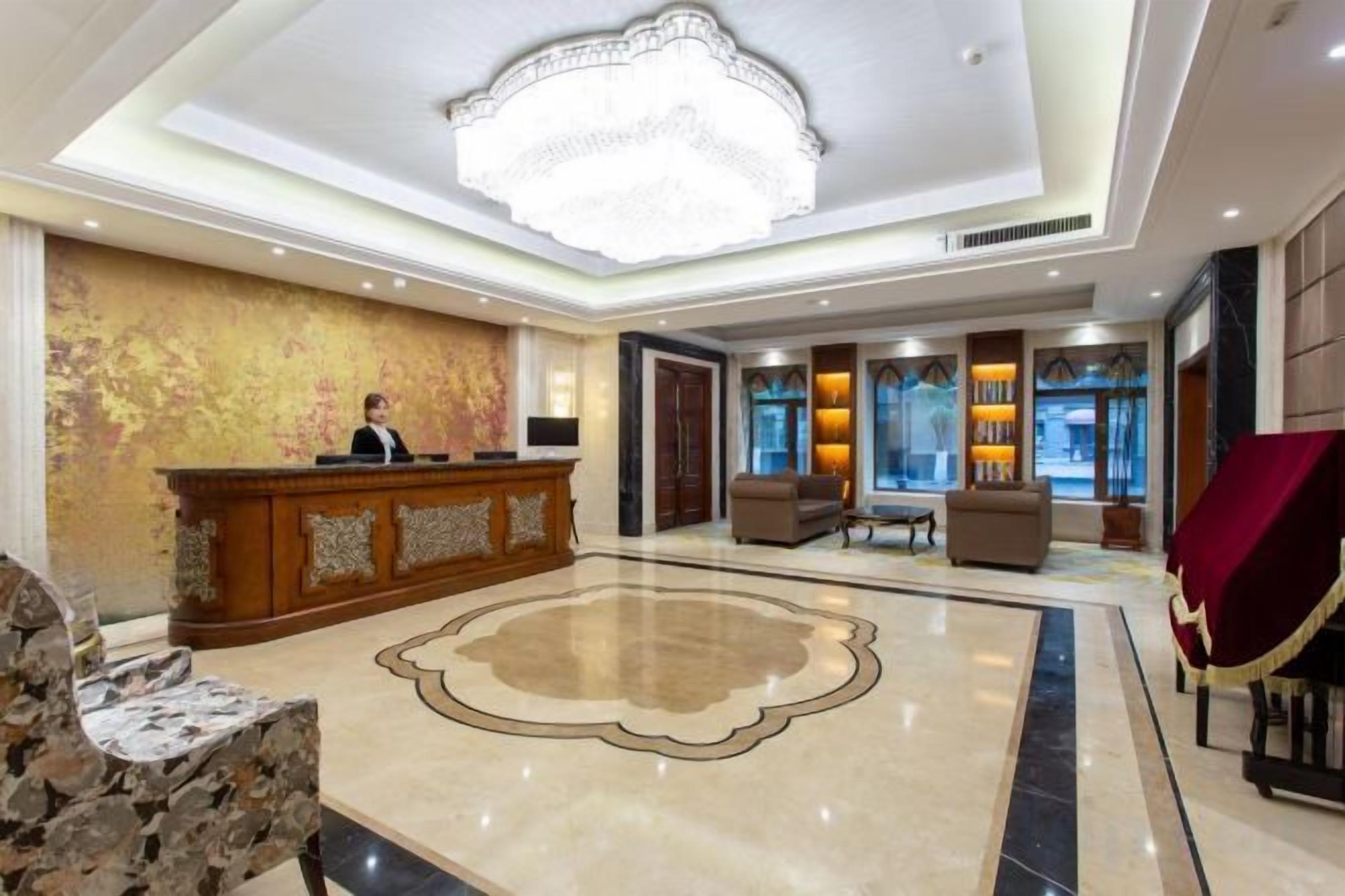 A hotel lobby with a reception desk, a chandelier, a large decorative rug, and seating areas.