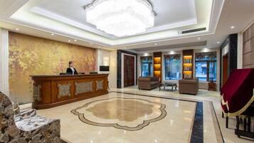 A hotel lobby with a reception desk, a chandelier, a large decorative rug, and seating areas.