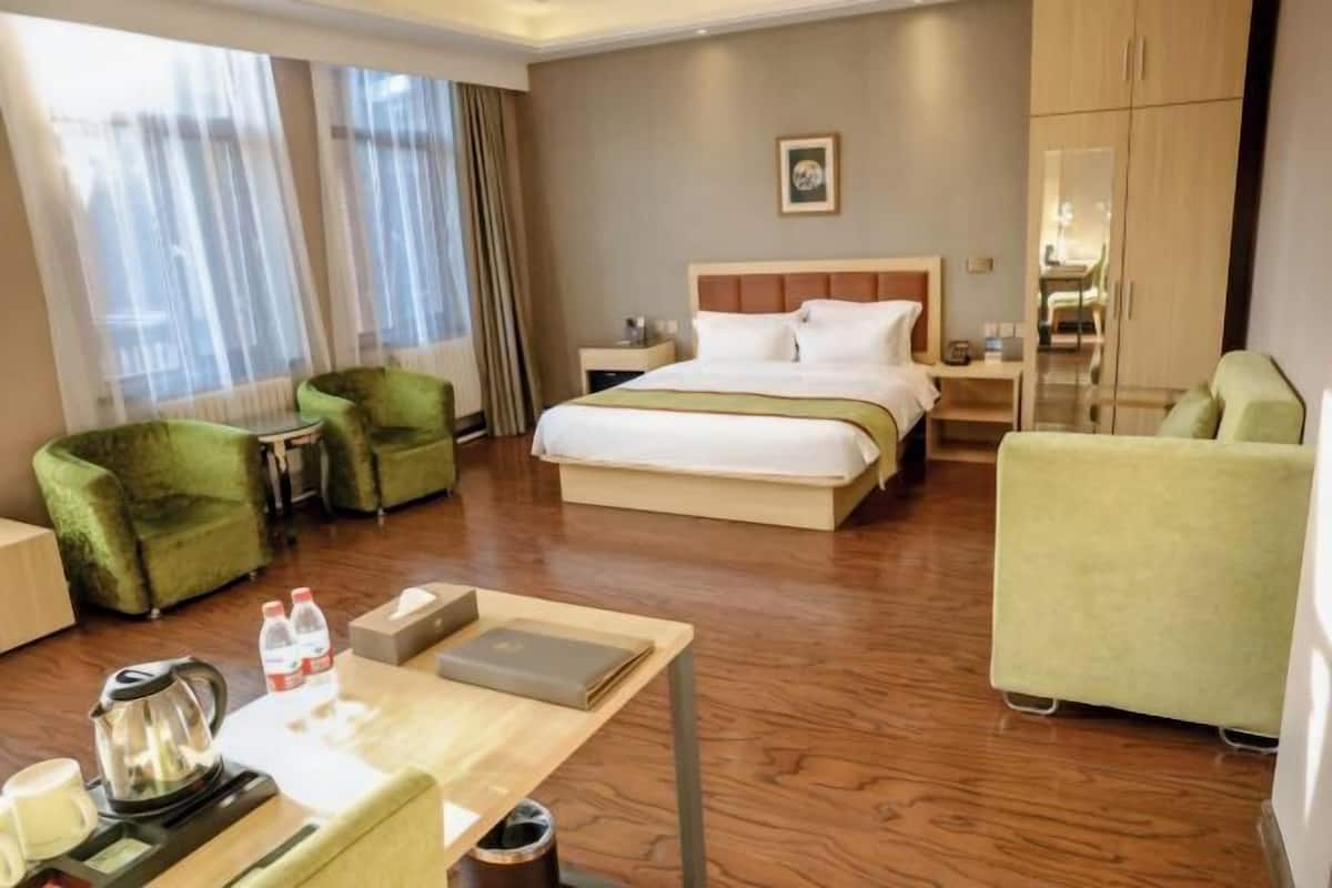 Une chambre d’hôtel avec un lit, deux fauteuils verts, une table en bois avec une bouilloire et un ordinateur portable, et une grande fenêtre avec des rideaux.