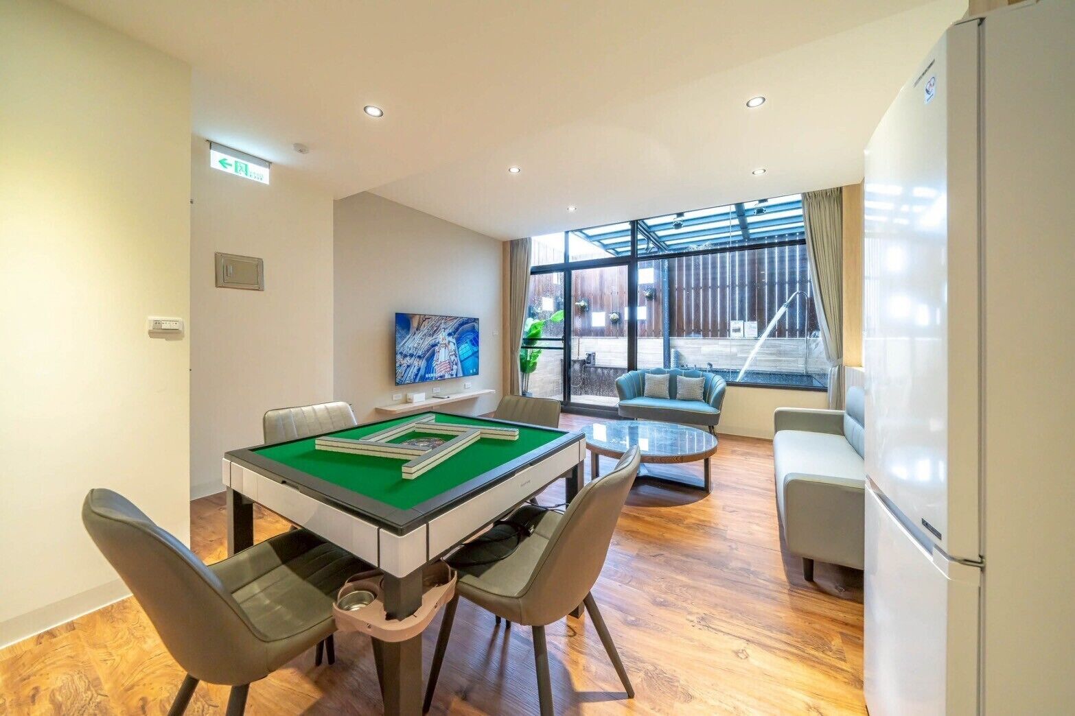 Une table de billard recouverte d’un drap vert, entourée de quatre chaises. Un canapé et une petite table sont placés près d’une grande fenêtre donnant sur l’extérieur.