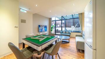 Une table de billard recouverte d’un drap vert, entourée de quatre chaises. Un canapé et une petite table sont placés près d’une grande fenêtre donnant sur l’extérieur.