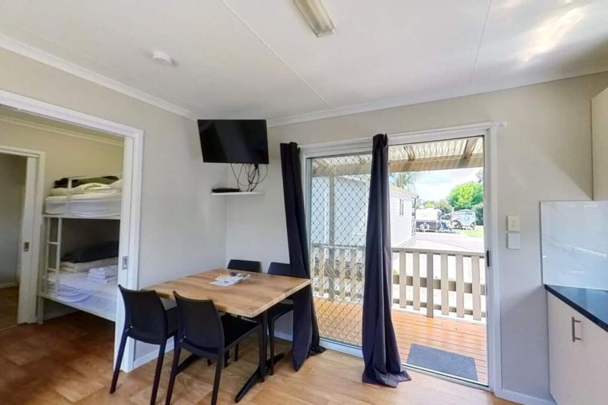 A compact room with a wooden dining table, black chairs, a TV mounted on the wall, and a sliding glass door leading to an outdoor deck.