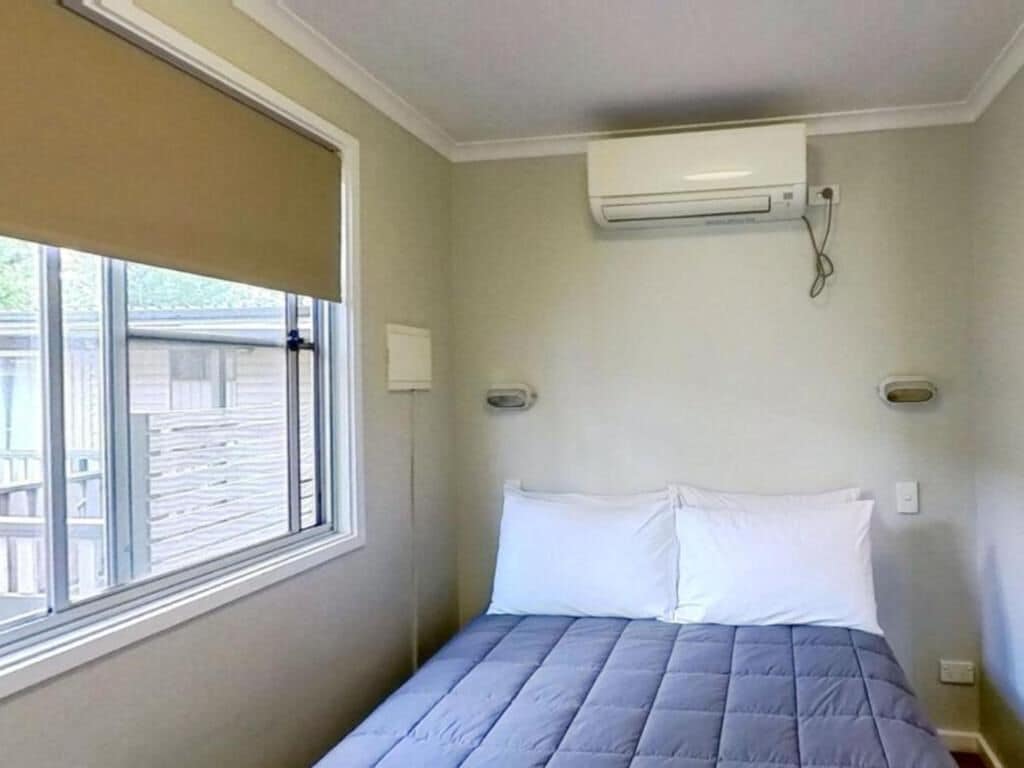 A bedroom with a bed, a window with blinds, an air conditioning unit, and a wall-mounted light fixture.