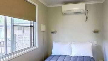 A bedroom with a bed, a window with blinds, an air conditioning unit, and a wall-mounted light fixture.